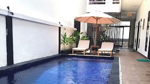 Una piscina con suelo de baldosas, rodeada por un muro blanco y una terraza de madera. Hay sillones de descanso y una pequeña mesa con sombrilla.