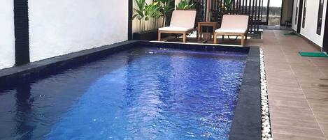 A swimming pool with tiled flooring, surrounded by a white wall and a wooden deck. There are lounge chairs and a small table with an umbrella.