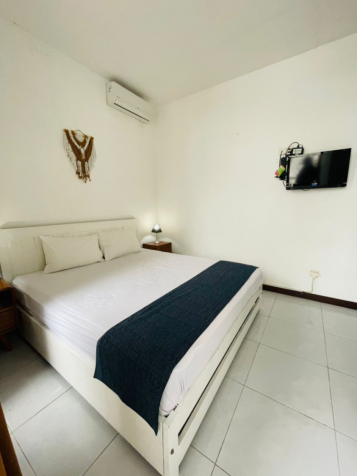 A neatly made bed with a dark blanket, a bedside lamp, a wall-mounted TV, and a decorative wall hanging.