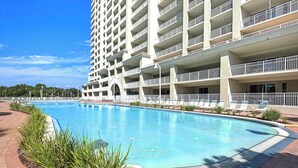 A large swimming pool surrounded by a landscaped area with plants, a multi-story building with balconies, and a clear blue sky.