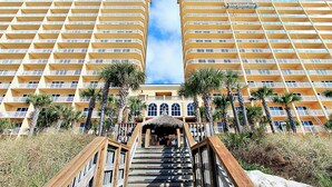 A hotel with a central staircase leading to a covered area, flanked by palm trees and a beach.