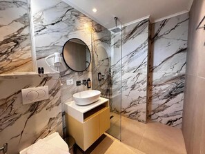 A modern bathroom with a marble backsplash, a round mirror, a white sink, and a glass shower enclosure. - Avenue Hotel (Ksamil)