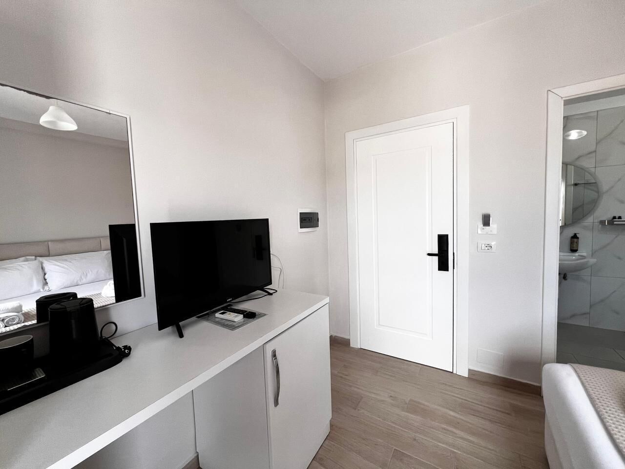 Una habitación de hotel moderna con un aparador blanco, un televisor de pantalla plana, una cafetera y una cama con cabecero blanco.