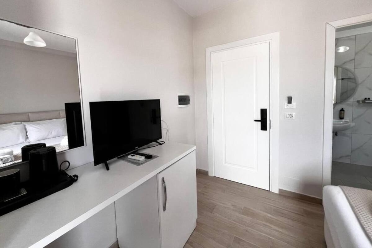 A modern hotel room with a flat-screen TV, a coffee machine, a white wardrobe, a white door, and a bathroom visible through a glass partition.