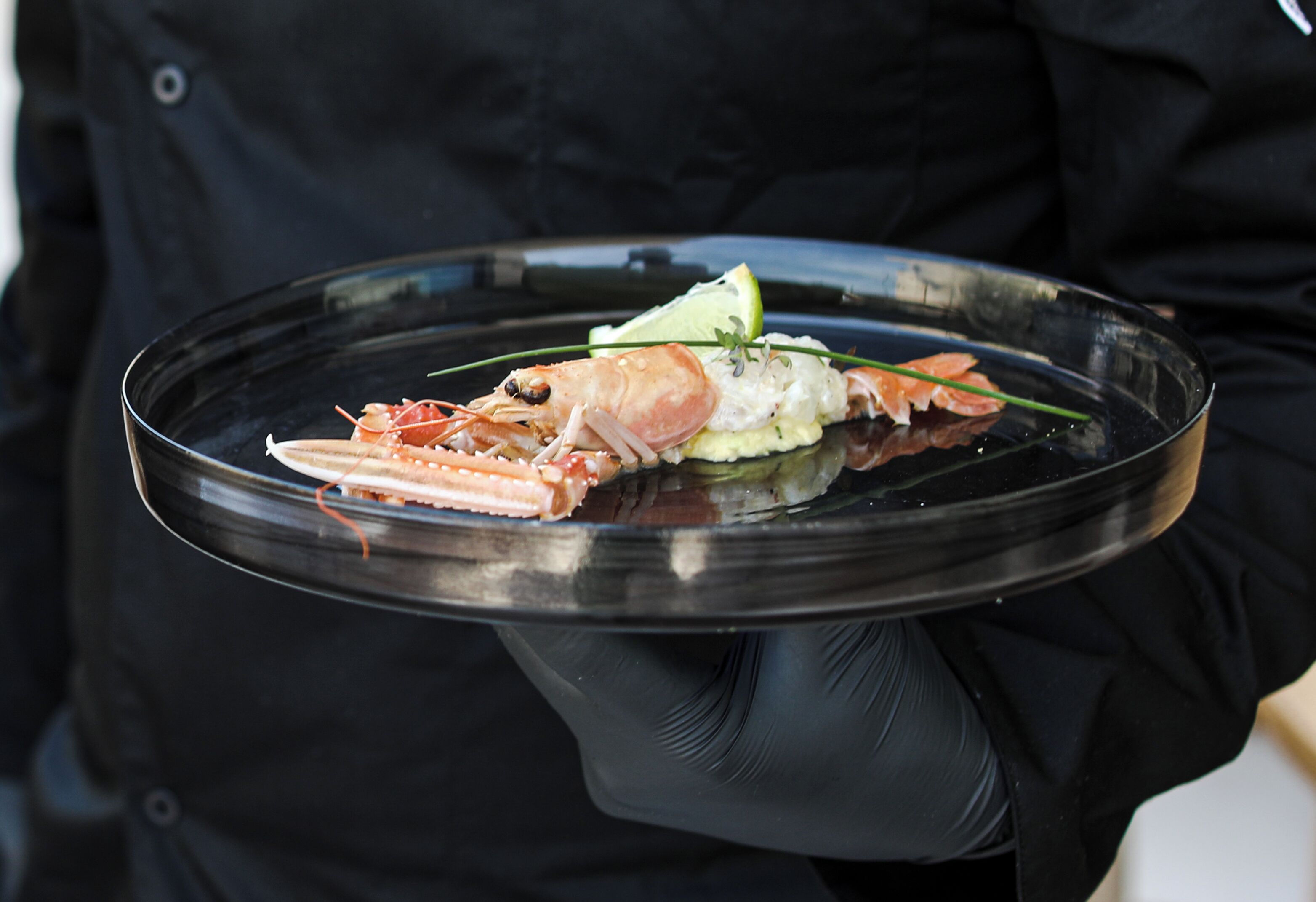 A person holding a clear glass plate with a lobster and garnishes.