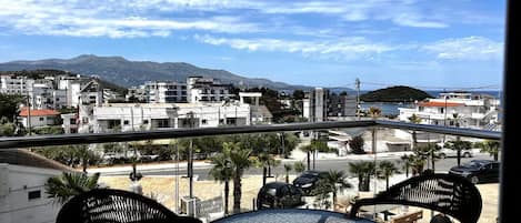 Un balcón con una mesa redonda y sillas, con vista a una zona costera con edificios y una montaña al fondo.