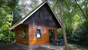 Una cabaña de madera con techo inclinado, base de ladrillo y un patio con mobiliario exterior.