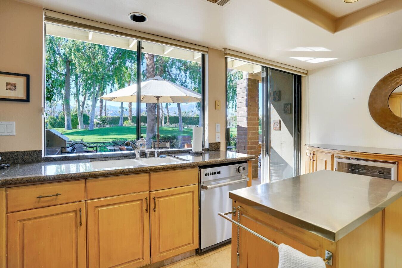 Une cuisine avec des meubles en bois, une cuisinière en acier inoxydable et une vue sur une cour arrière agrémentée d’un parasol.