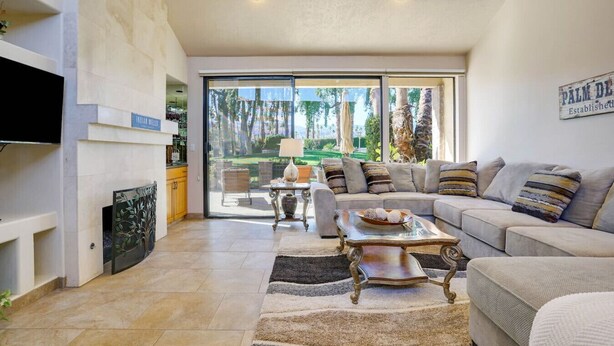 A spacious living room with a large sofa, a coffee table, and a TV mounted on the wall. There is a sliding glass door leading to an outdoor area with a view of trees. - Del Sol: Resort Style Living, BBQ, Fire Pit, Peace (Palm Desert)
