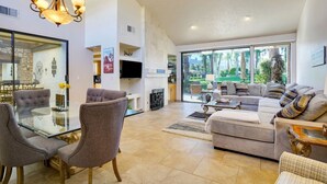 A spacious living room with a glass dining table, comfortable seating, a large window with a view of greenery, and a modern chandelier. - Del Sol: Resort Style Living, BBQ, Fire Pit, Peace (Palm Desert)