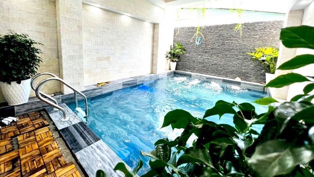 A swimming pool with a wooden deck area, surrounded by plants and a stone wall. - Ho Quy Ly Holiday Apartment for Family (Ho Chi Minh City)