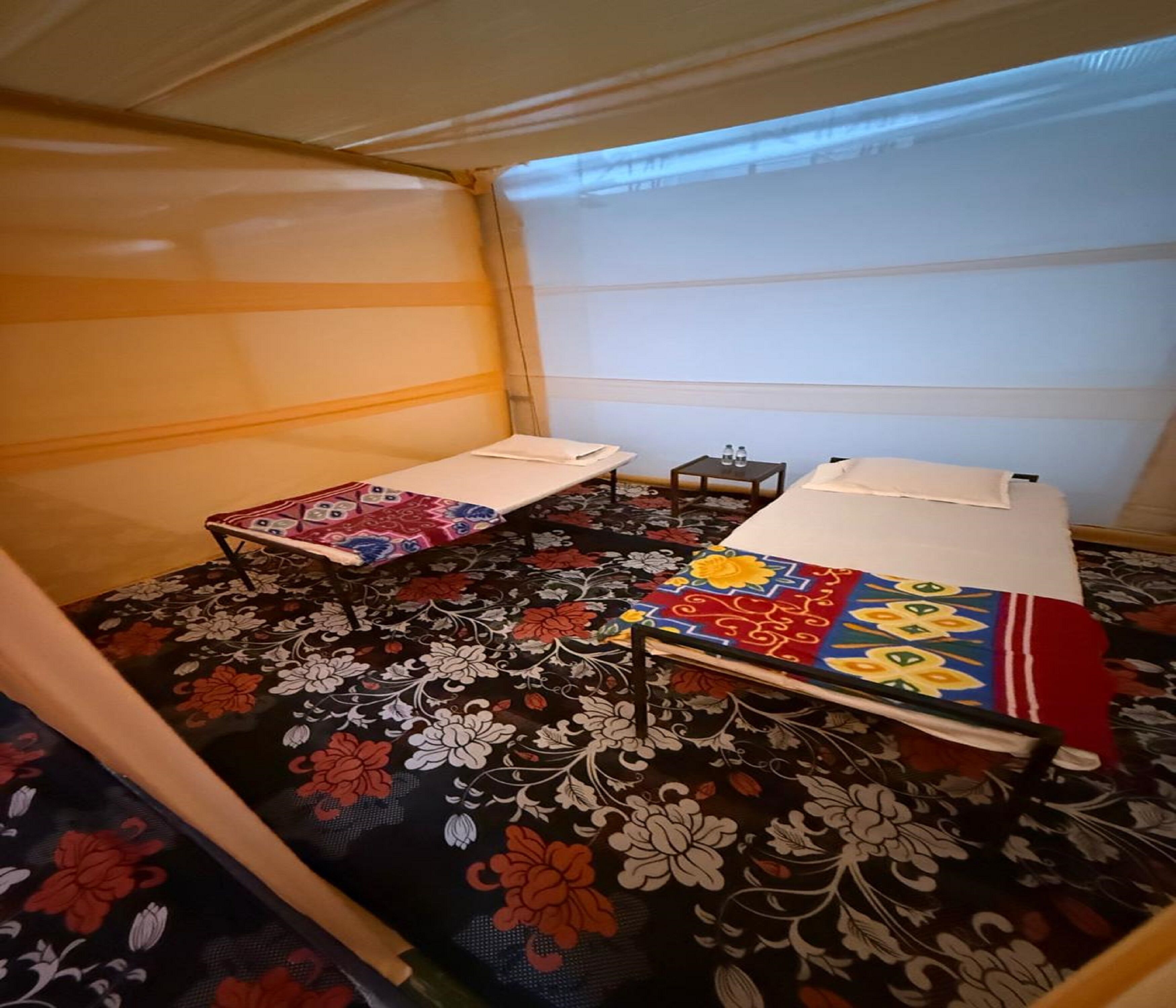 A room with two beds, each with a colorful bedspread. There is a small table with two bottles on it.