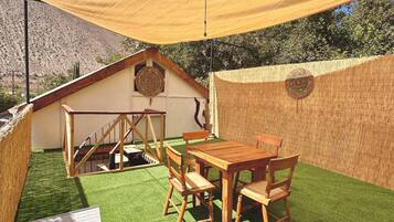 Relax under the shaded patio with a wooden table and chairs, surrounded by lush artificial grass and a serene bamboo fence.