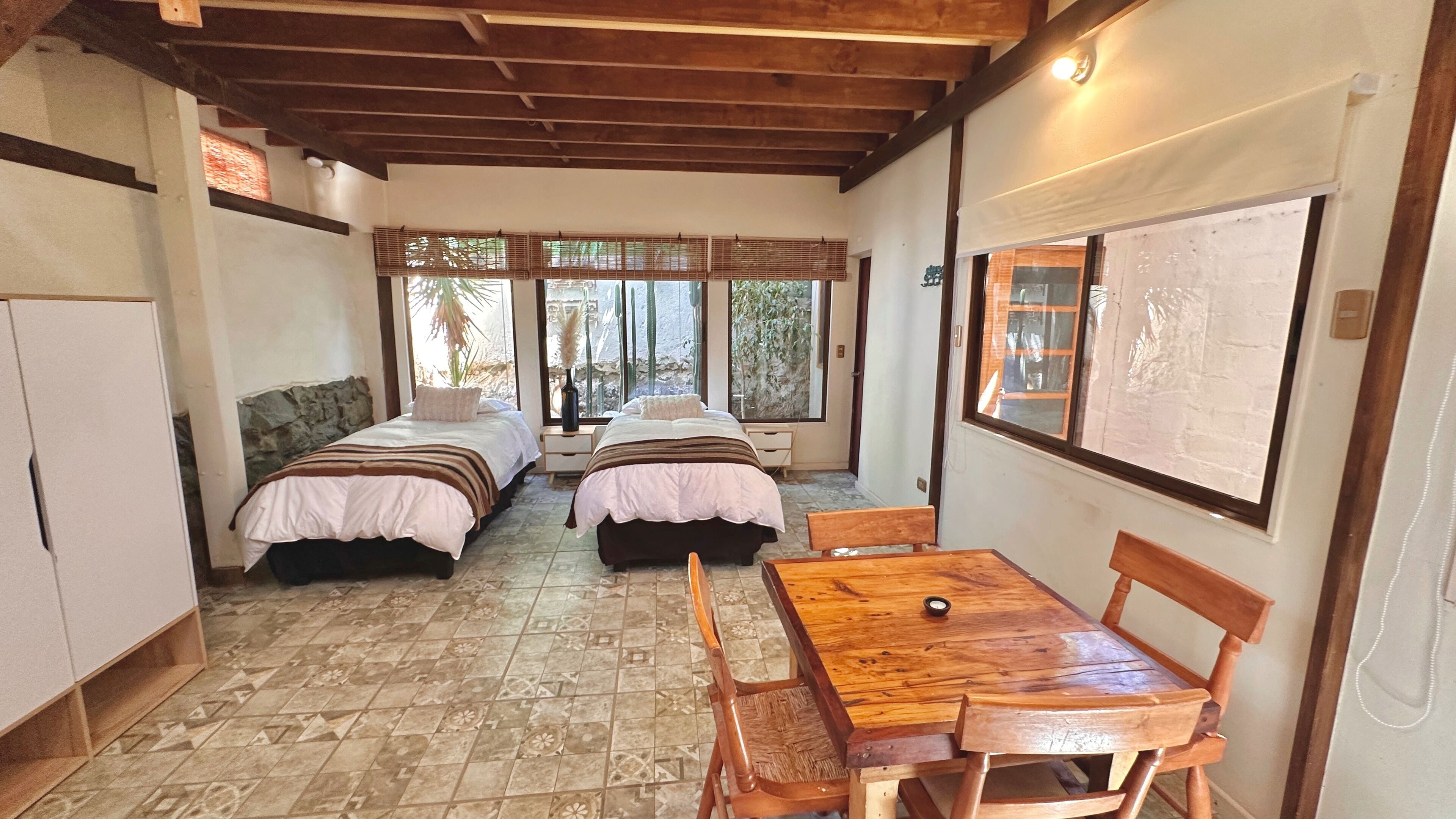 A room with two beds, a wooden table, and a view of the outdoors.