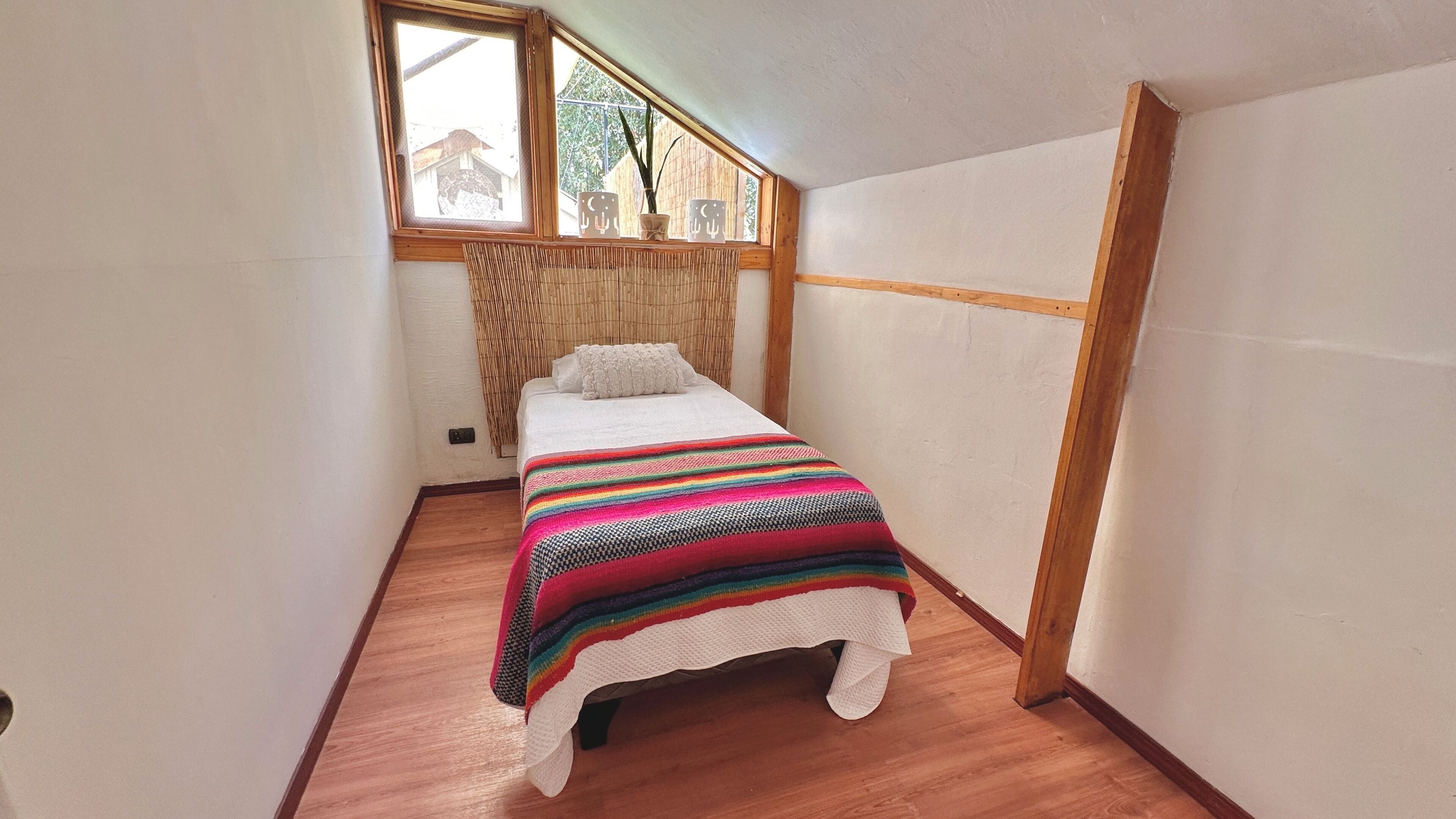 Una habitación pequeña y estrecha con una cama individual, una ventana que da a una casa y una cortina de bambú.