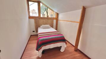 Una habitación pequeña y estrecha con una cama individual, una ventana que da a una casa y una cortina de bambú.