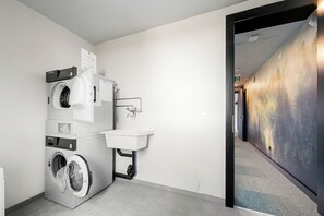 Une buanderie équipée d’une laveuse et d’une sécheuse superposées, d’un évier et d’un couloir menant à une pièce dont le mur est recouvert d’un revêtement texturé.