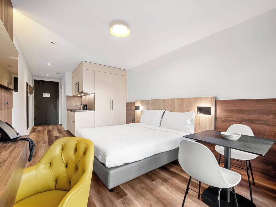 A modern hotel room with a bed, a desk, a chair, a telephone, and a wooden panel wall.