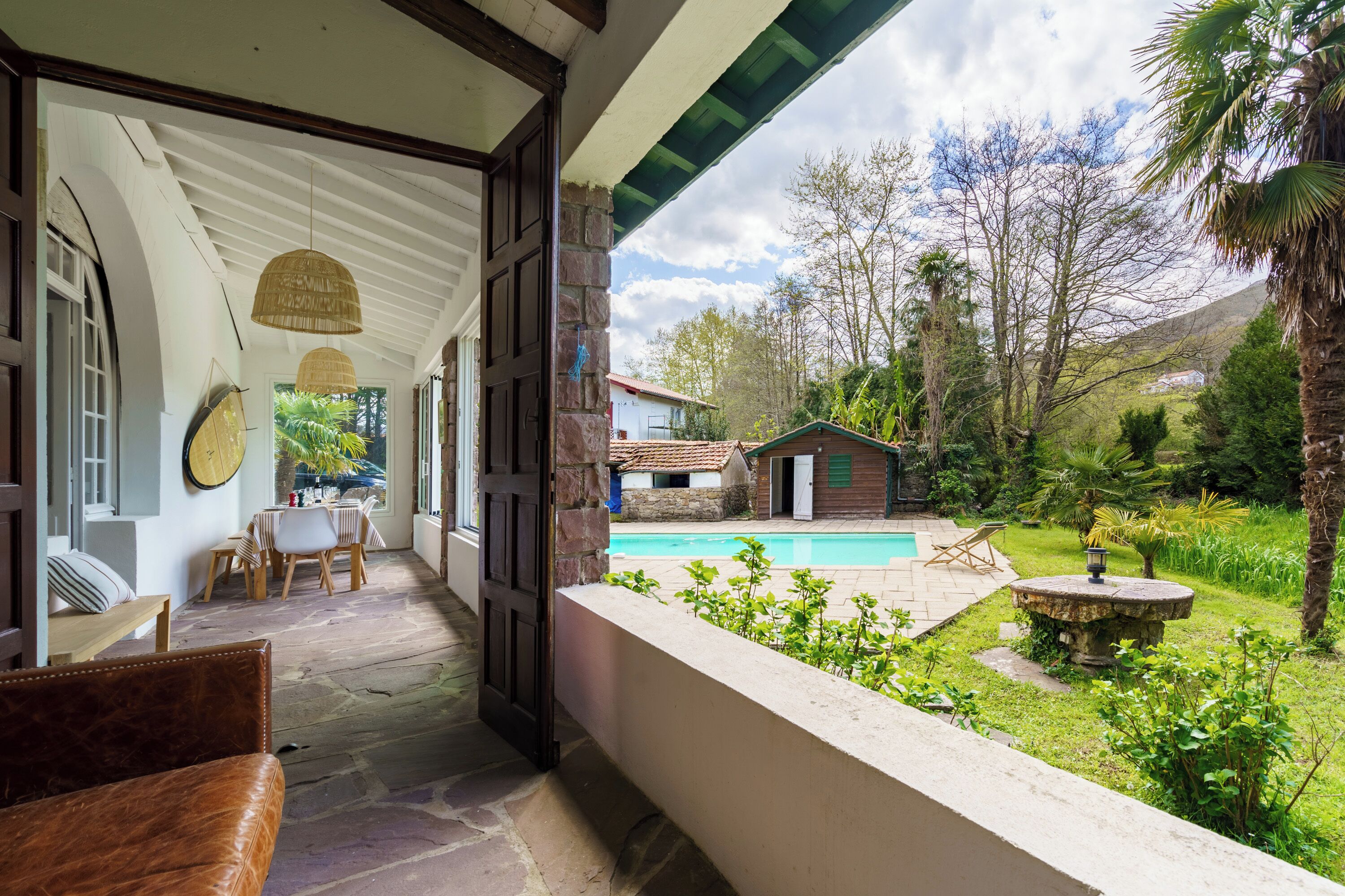 A covered outdoor area with a pool, stone flooring, and a view of a house and greenery.