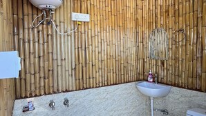 Baño con techo de madera, paneles de bambú en la pared, un lavamanos blanco, inodoro y zona de ducha con un taburete azul.