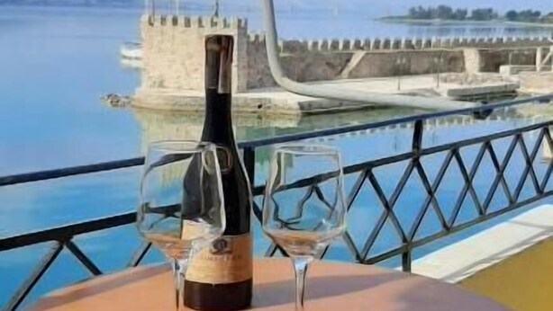 A table with a bottle and two glasses on a balcony overlooking a body of water and a historic structure.