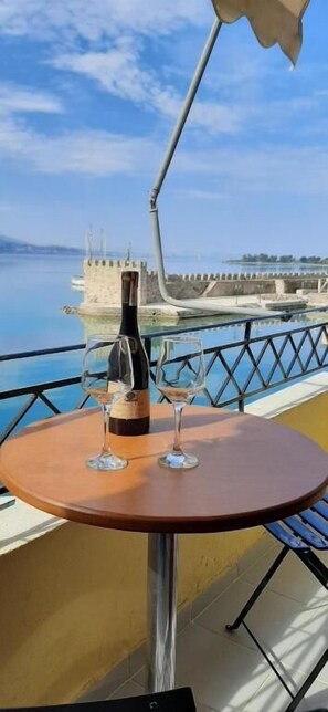 A table with a bottle and two glasses on a balcony overlooking a body of water and a historic structure. - Dr. J. Spon Apartment (Nafpaktos)