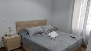 A neatly made bed with a grey blanket and pillows, a bedside lamp, and a small wooden nightstand with drawers.