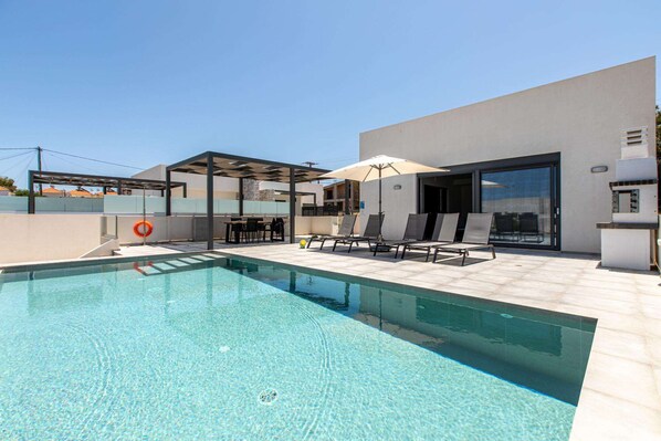 A modern outdoor pool area with clear blue water, lounge chairs, and a covered seating area.