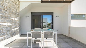 A modern outdoor patio with a glass table and chairs, a stone wall, and a sliding glass door leading to a building. - Menta Villa - Two Bedroom Villa, Sleeps 4 (Tavronitis)