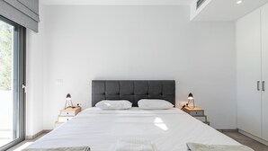 A modern bedroom with a large bed, two bedside tables with lamps, and a view of the outdoors through a large window. - Menta Villa - Two Bedroom Villa, Sleeps 4 (Tavronitis)