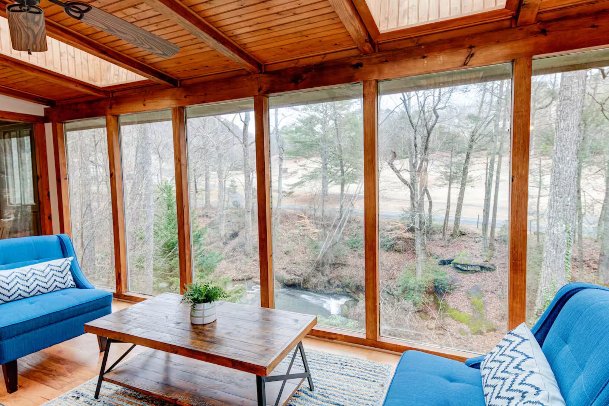 Ein Zimmer mit großem Fenster, das einen Blick auf Wald und Bach freigibt, mit einem blauen Sofa und einem Holztisch.
