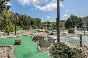 Um campo de mini golfe com superfície de putting verde, um obstáculo de pedra, um lago com uma fonte e uma ponte de madeira.