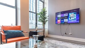 Una sala moderna con un televisor de pantalla plana grande montado en la pared, una mesa de centro de vidrio, un sofá naranja confortable y un ventanal con vistas a la ciudad.