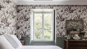 A bedroom with floral wallpaper, a large window, a bed with white bedding, a dresser, and a mirror with a floral arrangement. - Deluxe double room (Le Pian-Médoc)