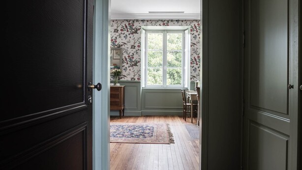 A room with a wooden floor, a window with floral wallpaper, a door, and a small table with a vase. - Deluxe double room (Le Pian-Médoc)