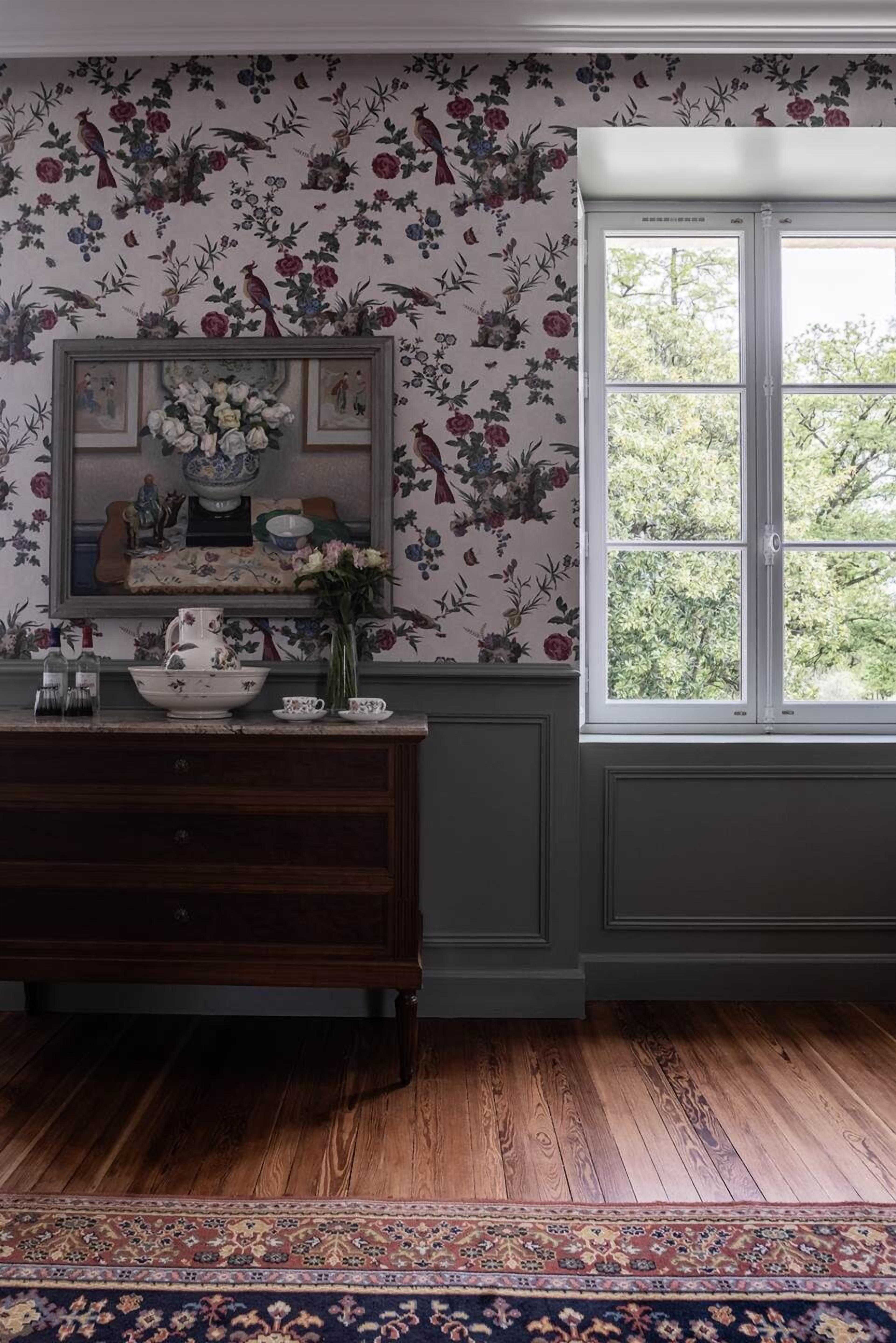 A room with floral wallpaper, a wooden dresser, a vase with flowers, a framed picture, and a window with a view of trees.