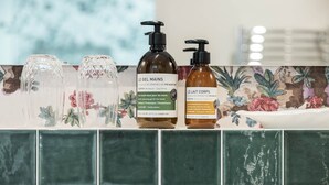 A bathroom shelf with two bottles of Le Gel Mains and Le Lait Corps body care products, and two clear glass bottles. - Deluxe double room (Le Pian-Médoc)