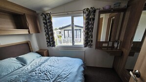 Ein Schlafzimmer mit Bett, Vorhängen, einem Fenster, einem Heizkörper, einem hölzernen Kopfteil, einem Nachttisch und einem Regal mit Gegenständen.