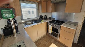 Eine kompakte Küche mit Spüle, Herd und Backofen. Ein Fenster mit Jalousien, ein Erste-Hilfe-Set und ein Wasserkocher sind vorhanden.
