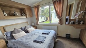 A bedroom with a bed, bedside tables, a window with curtains, a radiator, a small shelf with decorative items, and a bucket on the floor. - Heidi 2 Bed Robin Hood (Rhyl)