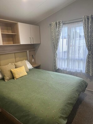 A bedroom with a bed, a wardrobe, a bedside table, and a window with curtains. - Mikey Caravan With Balcony (Rhyl)