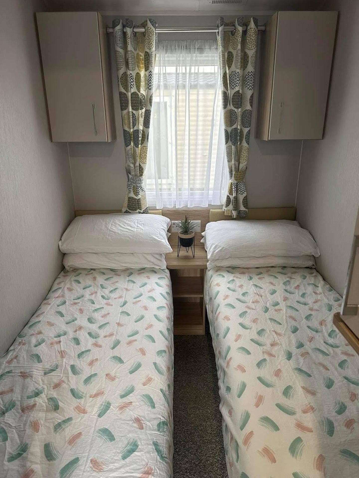 A small, compact room with two single beds, a small table, and a window with patterned curtains.