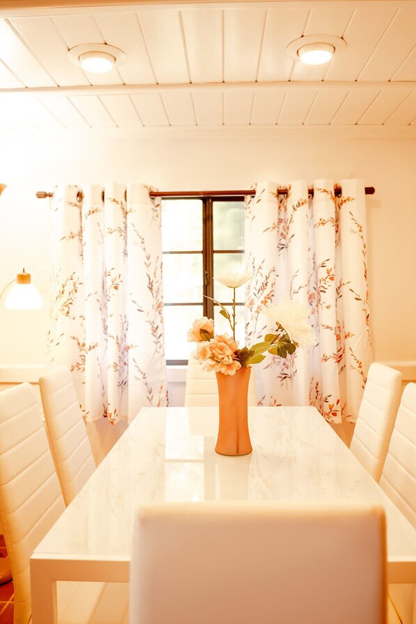 A dining area with a white table, floral curtains, and a vase of flowers.
