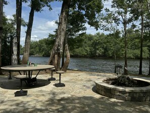 An outdoor patio with a round table and benches, surrounded by trees and a body of water.