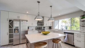A modern kitchen with white cabinets, stainless steel appliances, a central island with stools, pendant lighting, and a view of greenery outside. - Beryl Retreat (San Diego)