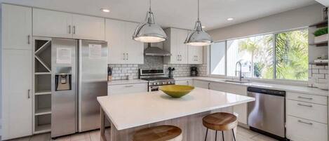 Una cocina moderna con armarios blancos, electrodomésticos de acero inoxidable, una isla central con taburetes, iluminación colgante y vistas a zonas verdes.