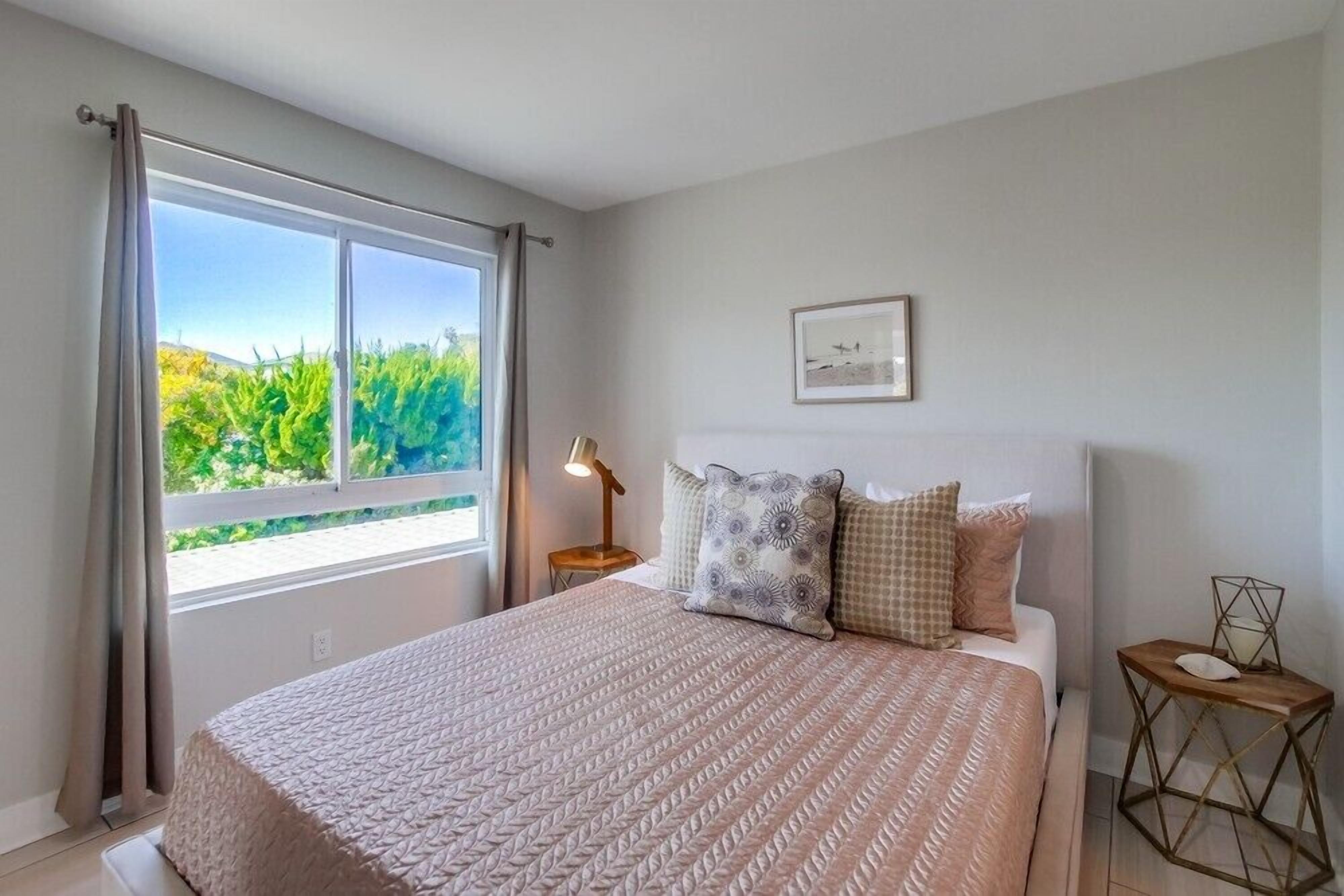 A bedroom with a large window, a bed with patterned bedding, a bedside lamp, and a framed picture on the wall.