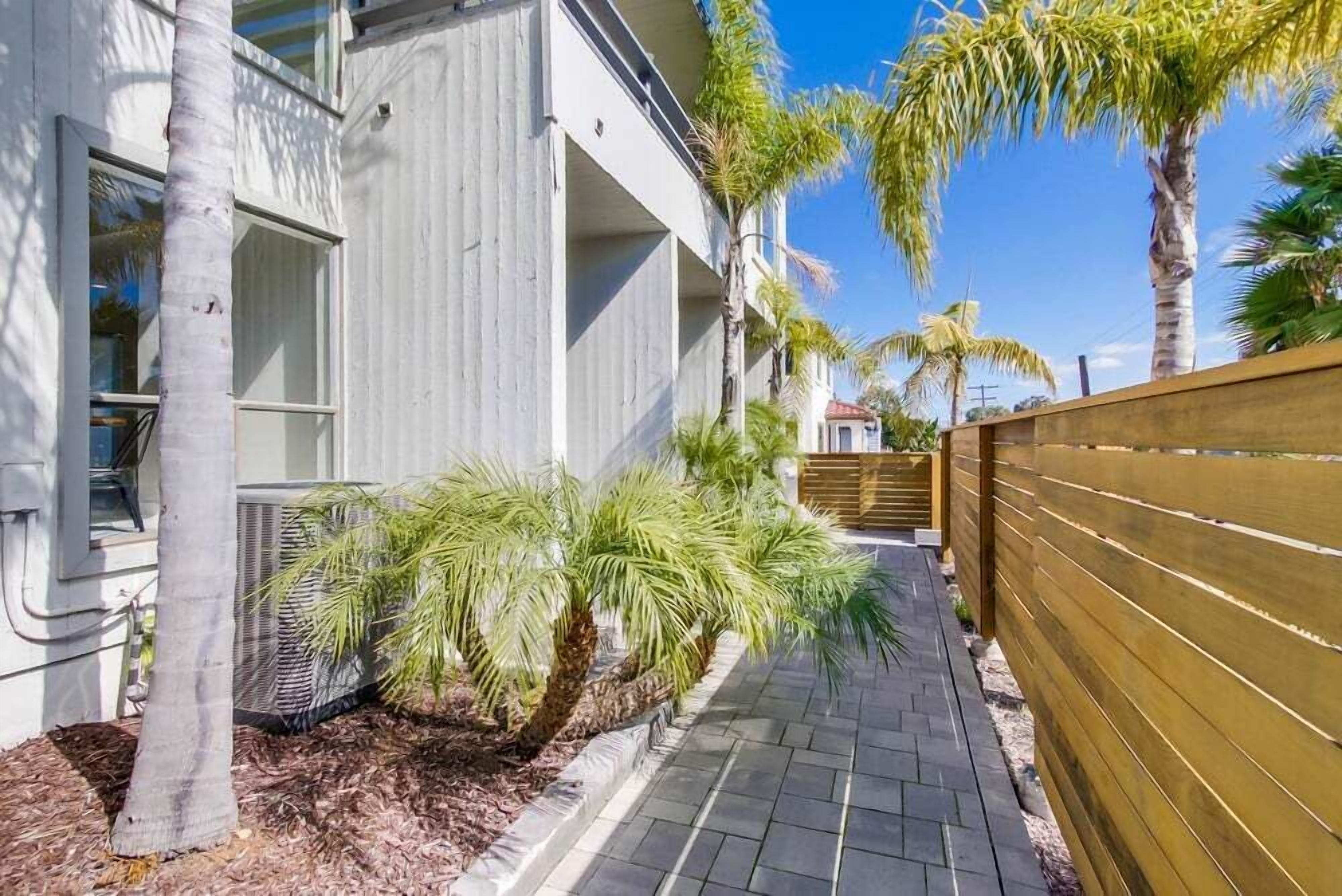 A pathway lined with palm trees and a wooden fence, leading to a building with large windows and an air conditioning unit.