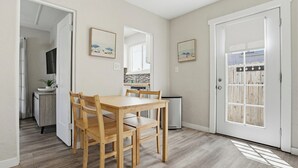 Een eethoek met een houten tafel en stoelen, een keuken op de achtergrond en een deur met een raam.