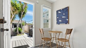 Een kamer met een deur die leidt naar een terras, een kleine ronde tafel met stoelen, een raam met jaloezieën en een abstract kunstwerk aan de muur.